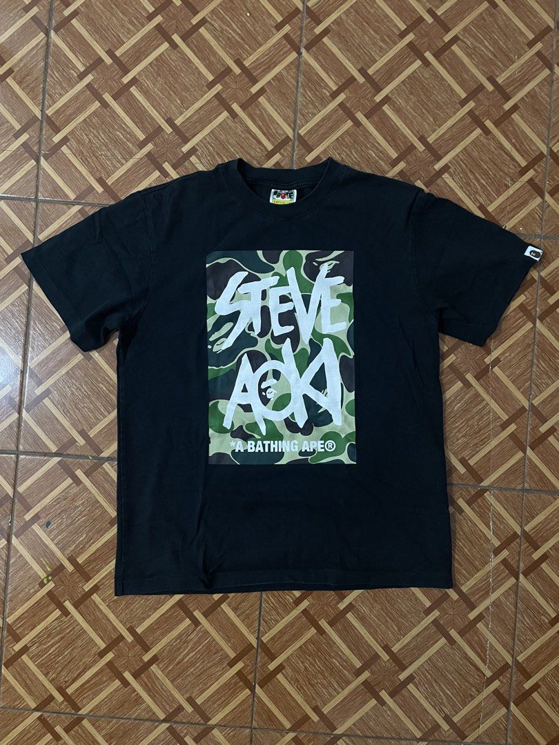 A Bathing Ape Steve Aoki Camo T-shirt (Category: Tshirts & Polo Shirts ) located in Manila. Submitted by ch1lleroli82692 (Listing ID 1466505156, Image 1). Description: - Brand: A Bathing Ape (BAPE) x Steve Aoki - Color: Black with green camo graphic - Design: Large Steve Aoki chest print, iconic ape head logo tag on left sleeve - Fit: Short sleeves, crew neck, solid black back - Condition: Pristine, well-kept streetwear - Shipping: Fast ship via 
