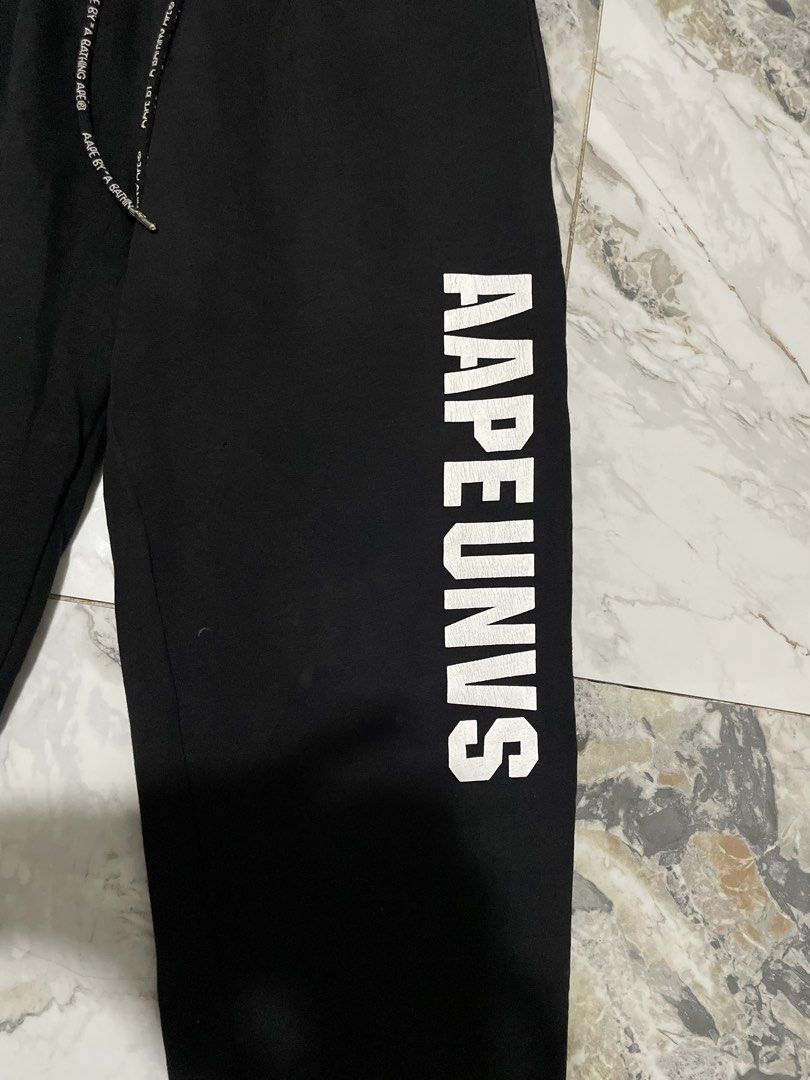 AAPE BY A BATHING APE® UNVS Moonface Logo Sweatpants (Category: Joggers) located in Antipolo. Submitted by divineyyy69 (Listing ID 1466525180, Image 2). Description: Authentic AAPE BY A BATHING APE® UNVS graphic joggers/sweatpants in black. Features the distinct vertical AAPEUNVS varsity-style text block on the left leg, a front circular rubberized moonface badge, and customized branded premium metal aglets. The back pocket sports an oversized ic