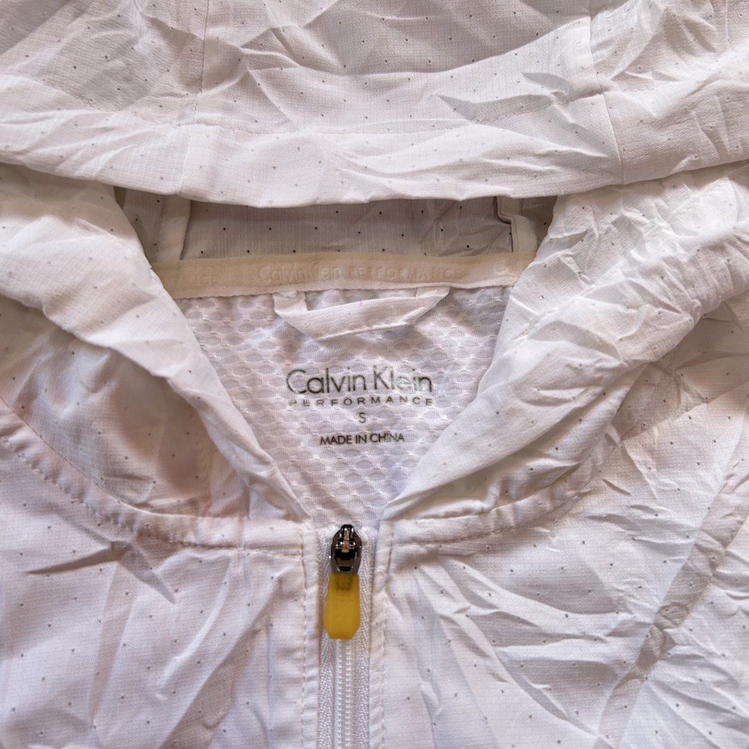 Calvin Klein Performance Windbreaker Jacket 🔥 (Category: Coats, Jackets and Outerwear) located in Jinoba-an. Submitted by eegiboy (Listing ID 1466426631, Image 2). Description: - Brand: Calvin Klein Performance - Color: White with bright orange sleeve accents - Size: Small dimes: 18/23 - Material: Lightweight perforated fabric for breathability - Design: Front zip closure with a pop of yellow zipper pull, hooded style - Branding: Calvin Klein lo