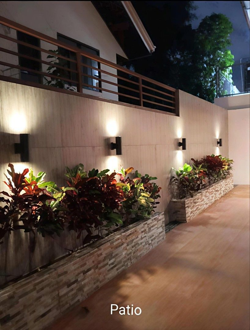 House For Sale (Category: House & Lot) located in Las Pinas. Submitted by mhay3188 (Listing ID 1466538472, Image 3). Description: Contemporary two-storey property. Includes a paved carport accommodating two vehicles, outdoor patio with landscaped planters, and ambient wall lighting. Interior spaces feature warm wood paneling, recessed ceiling lights, tiled floors, and an open layout dining area with built-in cabinetry. Clean finishes and modern a