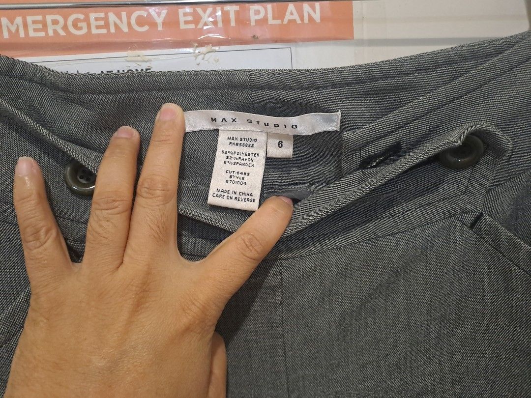 Max Studio gray pants wide leg (Category: Other Bottoms) located in Manila. Submitted by joanbalde39643 (Listing ID 1466440471, Image 2). Description: The image shows a max studio pair of grey wide-leg dress trousers hanging on a wooden hanger against a door. They feature an elegant high-waisted yoke waistband accented with asymmetric side button closures and unique curved, stitched front slash pockets..