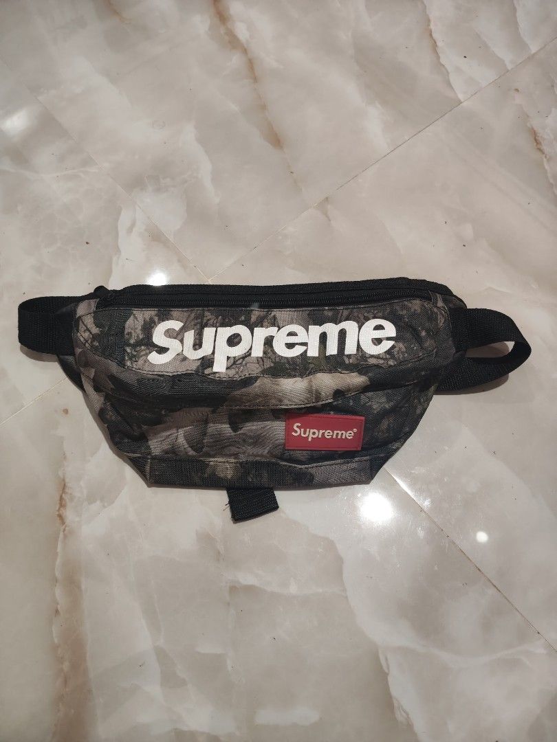 Supreme Camo Waist Bag (Category: Belt bags, Clutches and Pouches) located in Rizal, Mindoro Occidental. Submitted by finding_ukaygems5103 (Listing ID 1466509323, Image 1). Description: - Brand: Supreme - Style: Tree camouflage waist bag - Details: Large white Supreme lettering and iconic red box logo patch - Storage: Top zipper closure, ideal for phone, keys, and wallet - Fit: Adjustable black strap with durable plastic buckle - Condition: Good 