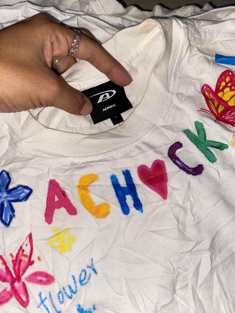 ACHOCK White Graphic Tee Hand-Drawn Design Size S (Category: Tshirts & Polo Shirts ) located in Muntinlupa City. Submitted by enajp82 (Listing ID 1466725199, Image 2). Description: - Brand: ACHOCK - Color: White - Size: Tagged S (fits S to M) - Design: Colourful hand-drawn style doodles (flowers, butterfly, palm tree, stick figures, and letter prints) - Fit: Relaxed fit, short sleeves - Style: Casual streetwear.