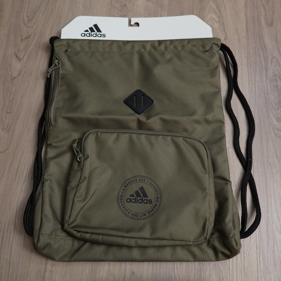 Adidas Olive Green Gym Sack Drawstring Bag (Category: Backpacks) located in Manila. Submitted by gentlewind_62f064 (Listing ID 1466744618, Image 1). Description: - Brand: Adidas - Color: Olive green with black rope drawstrings - Condition: Brand new with original paper tag - Pockets: Front zippered pocket with circular Adidas logo print and an additional vertical side zip pocket - Closure: Durable black rope drawstrings - Perfect for daily use, g