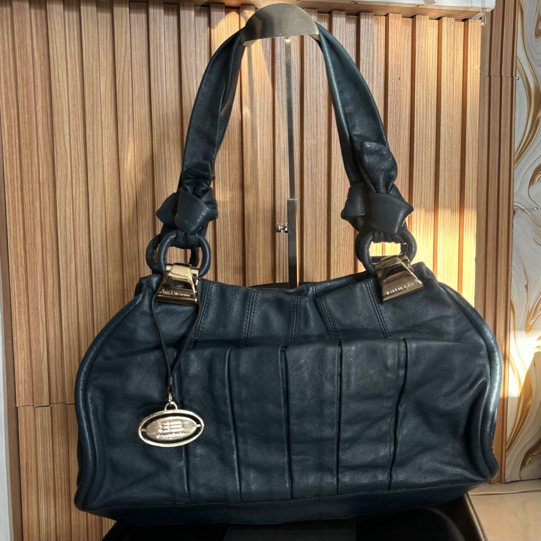 Balenciaga Blue Leather Shoulder Bag (Category: Luxury Bags & Wallets) located in Bambang. Submitted by heats777 (Listing ID 1466664076, Image 1). Description: - Brand: Balenciaga - Material: Premium dark blue leather - Hardware: Gold-tone with engraved logo plates and ring details - Design: Vertical pleat details on front/back with a hanging metal tag charm - Condition: Minor scuffs on the gold clasp and a small color rub spot on the bottom pipi