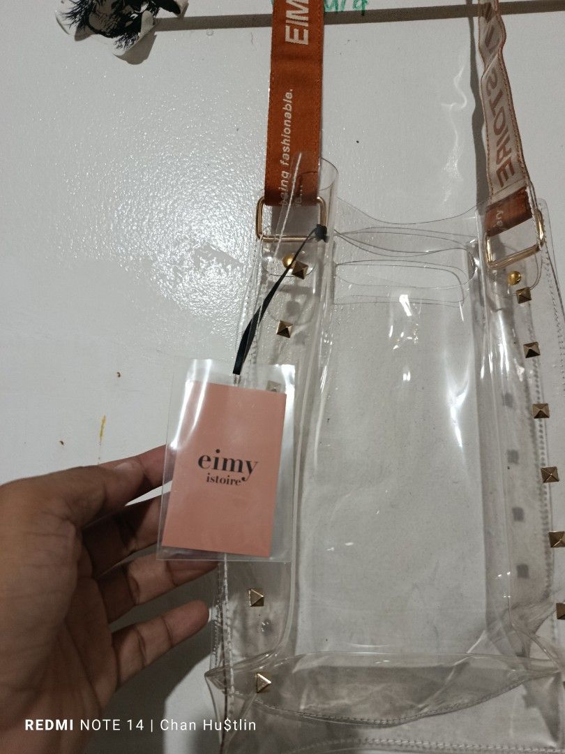 Eimy Istoire Clear Studded Shoulder Bag (Category: Shoulder Bags) located in Calamba. Submitted by chanmagpantay (Listing ID 1466590565, Image 3). Description: - Brand: Eimy Istoire (Authentic with original Japanese store tags) - Material: Durable clear PVC with a structured base - Accents: Chic gold pyramid studs along the sides - Strap: Bold orange fabric strap featuring vertical brand printing - Design: Open top for easy access - Condition: Br
