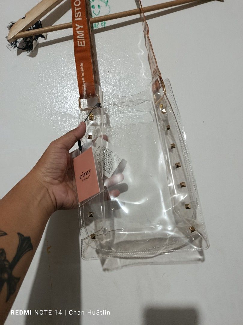 Eimy Istoire Clear Studded Shoulder Bag (Category: Shoulder Bags) located in Calamba. Submitted by chanmagpantay (Listing ID 1466590565, Image 2). Description: - Brand: Eimy Istoire (Authentic with original Japanese store tags) - Material: Durable clear PVC with a structured base - Accents: Chic gold pyramid studs along the sides - Strap: Bold orange fabric strap featuring vertical brand printing - Design: Open top for easy access - Condition: Br