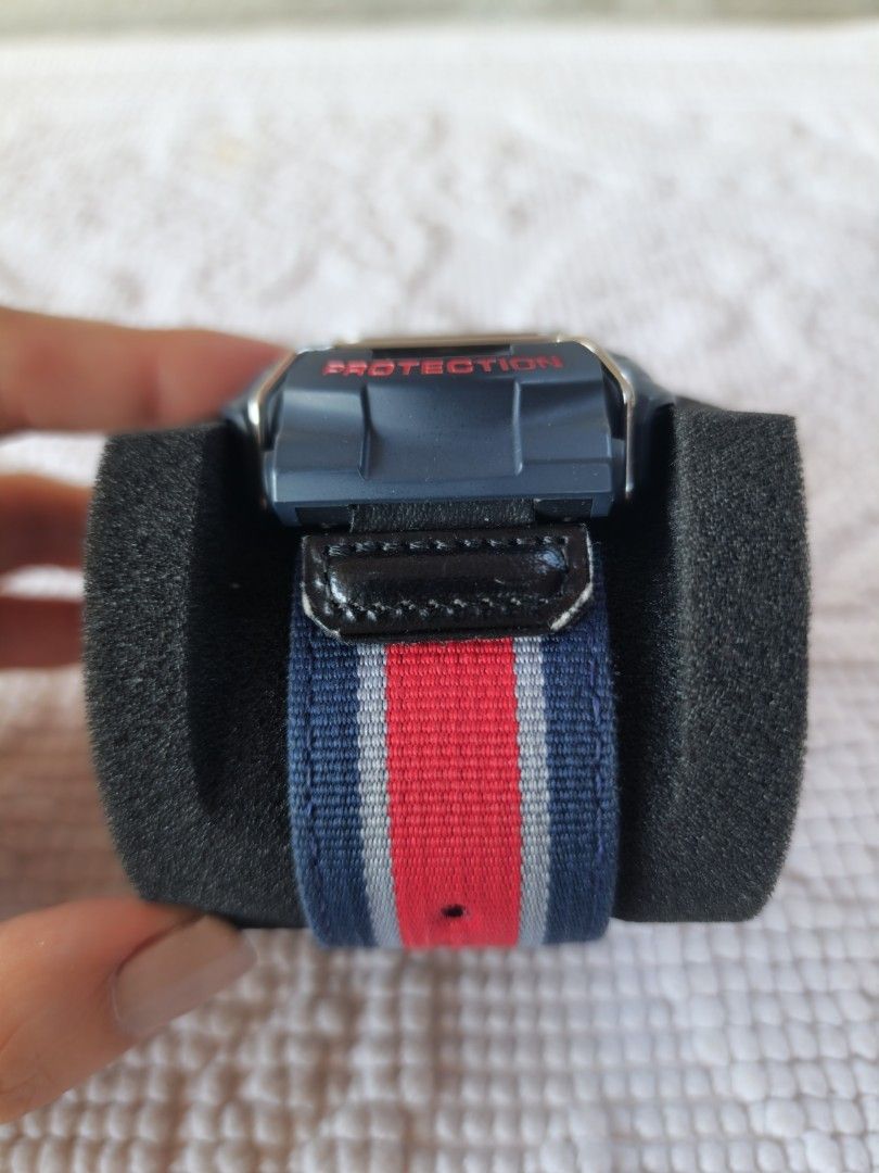 GShock (Category: Men's Watches) located in Imus. Submitted by alecsssss (Listing ID 1466565401, Image 3). Description: - Brand: Casio G-Shock GA 100 - Design: Blue case with protective metal wire guards over the dial - Strap: Red, white, and blue fabric strap with a secure silver metal buckle - Display: Dual analog and digital display face with bold red markings - Condition: Good pre-loved condition with a small scuff mark on the loop near the s