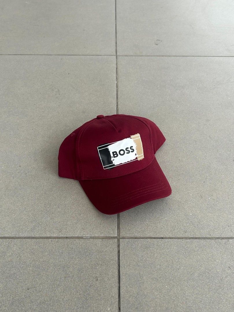 Hugo Boss Maroon Baseball Cap with Graphic Logo (Category: Caps & Hats) located in Pasig City. Submitted by paulclzdo (Listing ID 1466639270, Image 1). Description: Maroon baseball cap from Hugo Boss. Large graphic patch on the front with black, white, and gold brushstroke design and bold BOSS lettering. Adjustable strapback closure in the back with metal buckle. Inside has printed brand tags and taped seams. Clean condition overall, ready to wea