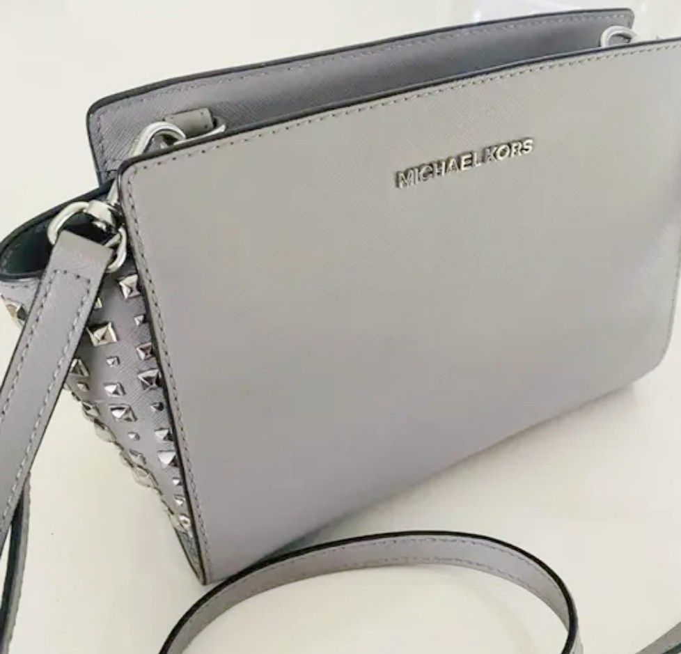 Michael Kors Grey Crossbody Bag with Studs (Category: Cross-body Bags) located in Manila. Submitted by lene48 (Listing ID 1466626931, Image 1). Description: Light grey Saffiano leather Michael Kors crossbody bag. Silver-tone hardware and stud details along the side gussets. Adjustable shoulder strap attached with metal clips. Zip top closure. Fabric lined interior includes one slip pocket and one zip pocket. Interior brand tag with QR code label 