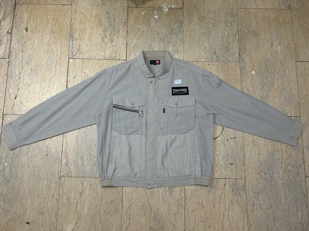 Thrasher Magazine Grey Workwear Jacket (Category: Coats, Jackets and Outerwear) located in Mandaluyong City. Submitted by 0lg2026 (Listing ID 1466751283, Image 1). Description: - Style: Grey work jacket with collar and full zip closure - Branding: Thrasher patch logo on the left chest and large stacked flame graphic on the back - Storage: Dual front button-flap chest pockets plus an extra right-side zip pocket - Fit & Comfort: Elasticated hem, bu