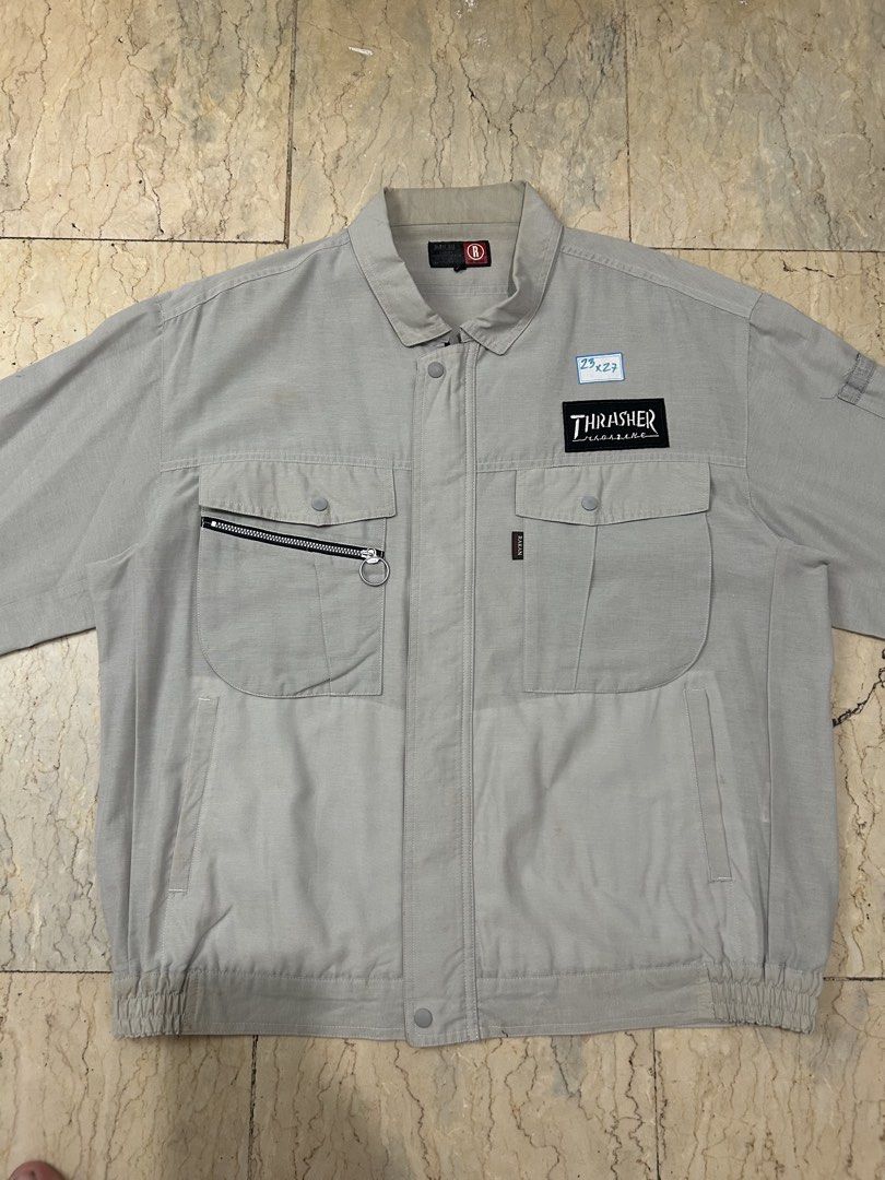 Thrasher Magazine Grey Workwear Jacket (Category: Coats, Jackets and Outerwear) located in Mandaluyong City. Submitted by 0lg2026 (Listing ID 1466751283, Image 2). Description: - Style: Grey work jacket with collar and full zip closure - Branding: Thrasher patch logo on the left chest and large stacked flame graphic on the back - Storage: Dual front button-flap chest pockets plus an extra right-side zip pocket - Fit & Comfort: Elasticated hem, bu