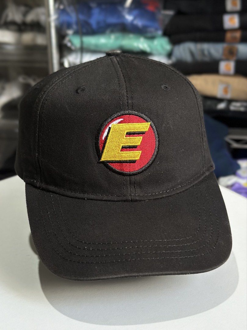 Black Cap with Embroidered E Logo (Category: Caps & Hats) located in Daet. Submitted by thrifthings2018 (Listing ID 1466867025, Image 2). Description: Black baseball cap, cotton material. Structured crown with embroidered 'E' patch logo on the front inside a red and black circle. Curved brim with stitching details. Adjustable snapback closure at the back. Clean condition overall, ready to wear..