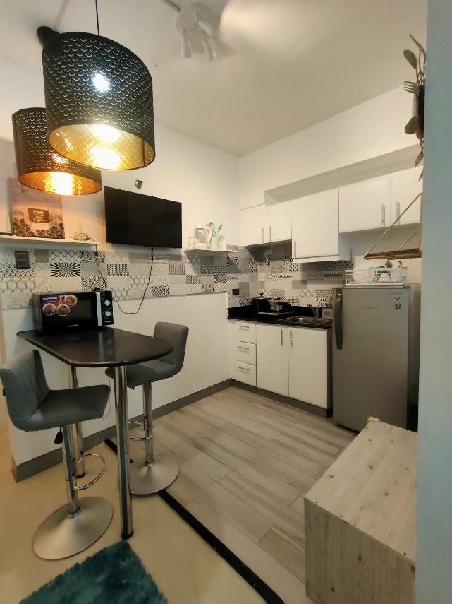 Fully furnished studio apartment interior setup with bed and dining (Category: Apartments & Condos) located in Makati City. Submitted by theacd (Listing ID 1466907671, Image 3). Description: - Complete studio apartment interior setup, ready for move-in - Double bed with stylish blue bedding and fluffy teal rug - Dining counter paired with two grey bar stools - Fully equipped kitchen area with overhead cabinets, refrigerator, and microwave - Elega