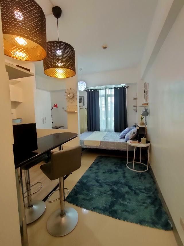 Fully furnished studio apartment interior setup with bed and dining (Category: Apartments & Condos) located in Makati City. Submitted by theacd (Listing ID 1466907671, Image 2). Description: - Complete studio apartment interior setup, ready for move-in - Double bed with stylish blue bedding and fluffy teal rug - Dining counter paired with two grey bar stools - Fully equipped kitchen area with overhead cabinets, refrigerator, and microwave - Elega