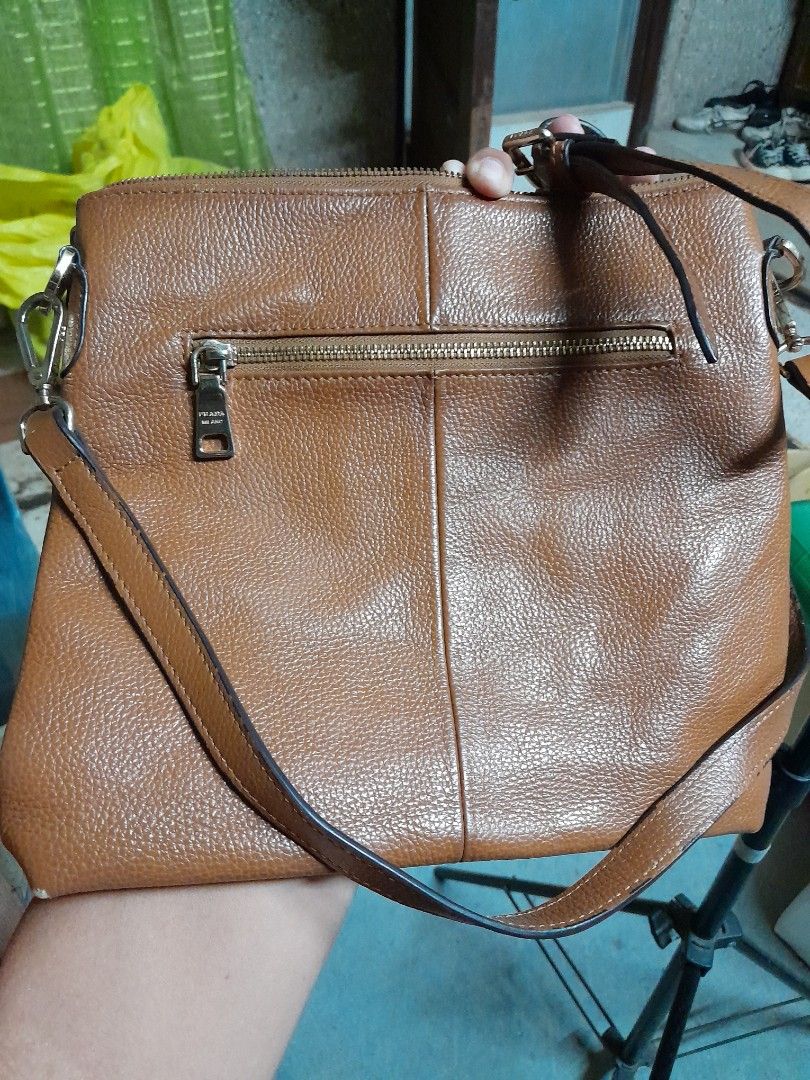 Prada Brown Leather Crossbody Bag (Category: Luxury Bags & Wallets) located in Manila. Submitted by olivertand55309 (Listing ID 1466906790, Image 2). Description: Tan brown pebbled leather crossbody bag from Prada. Debossed logo text on the front flap and metal brand plaque near the zipper pocket. Adjustable leather shoulder strap attached via metal hardware engraved with the brand name. Exterior zip pocket on both front and back panels. Gold-ton