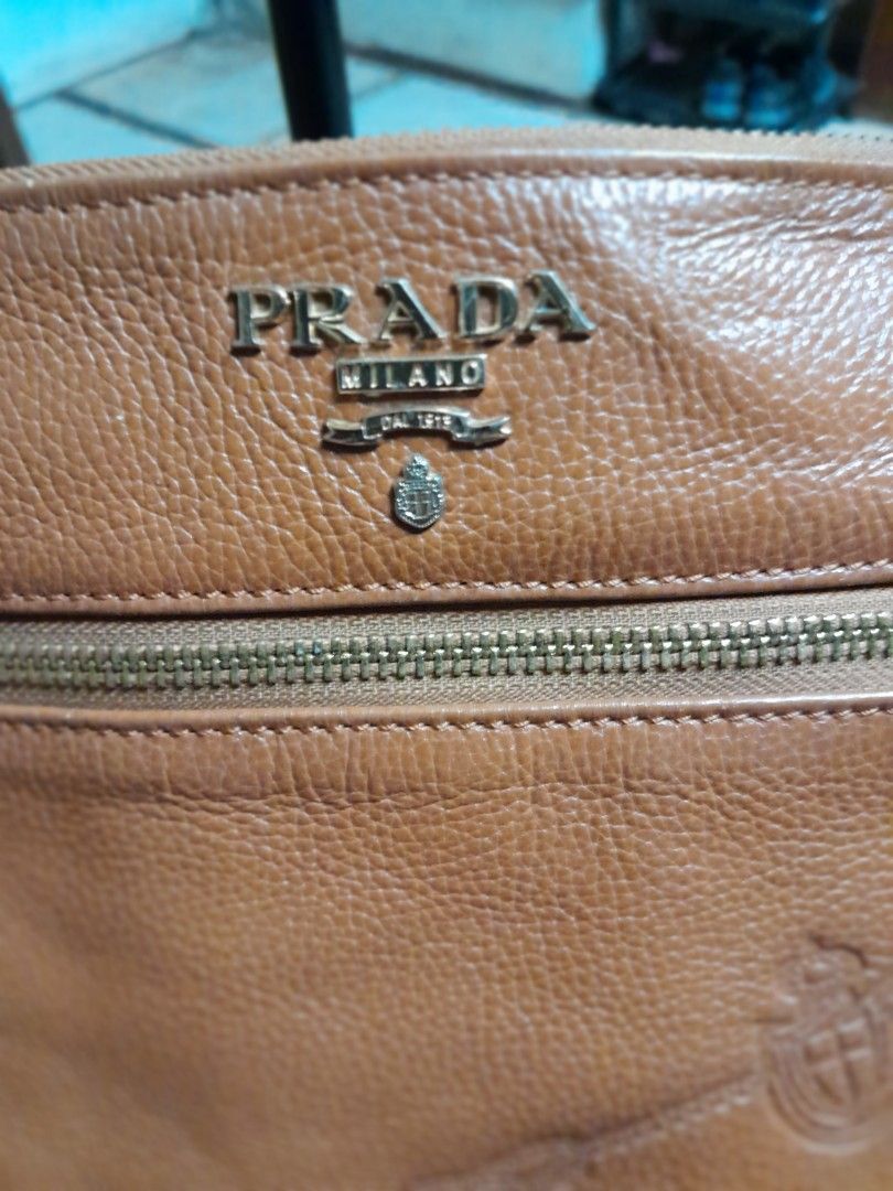 Prada Brown Leather Crossbody Bag (Category: Luxury Bags & Wallets) located in Manila. Submitted by olivertand55309 (Listing ID 1466906790, Image 3). Description: Tan brown pebbled leather crossbody bag from Prada. Debossed logo text on the front flap and metal brand plaque near the zipper pocket. Adjustable leather shoulder strap attached via metal hardware engraved with the brand name. Exterior zip pocket on both front and back panels. Gold-ton