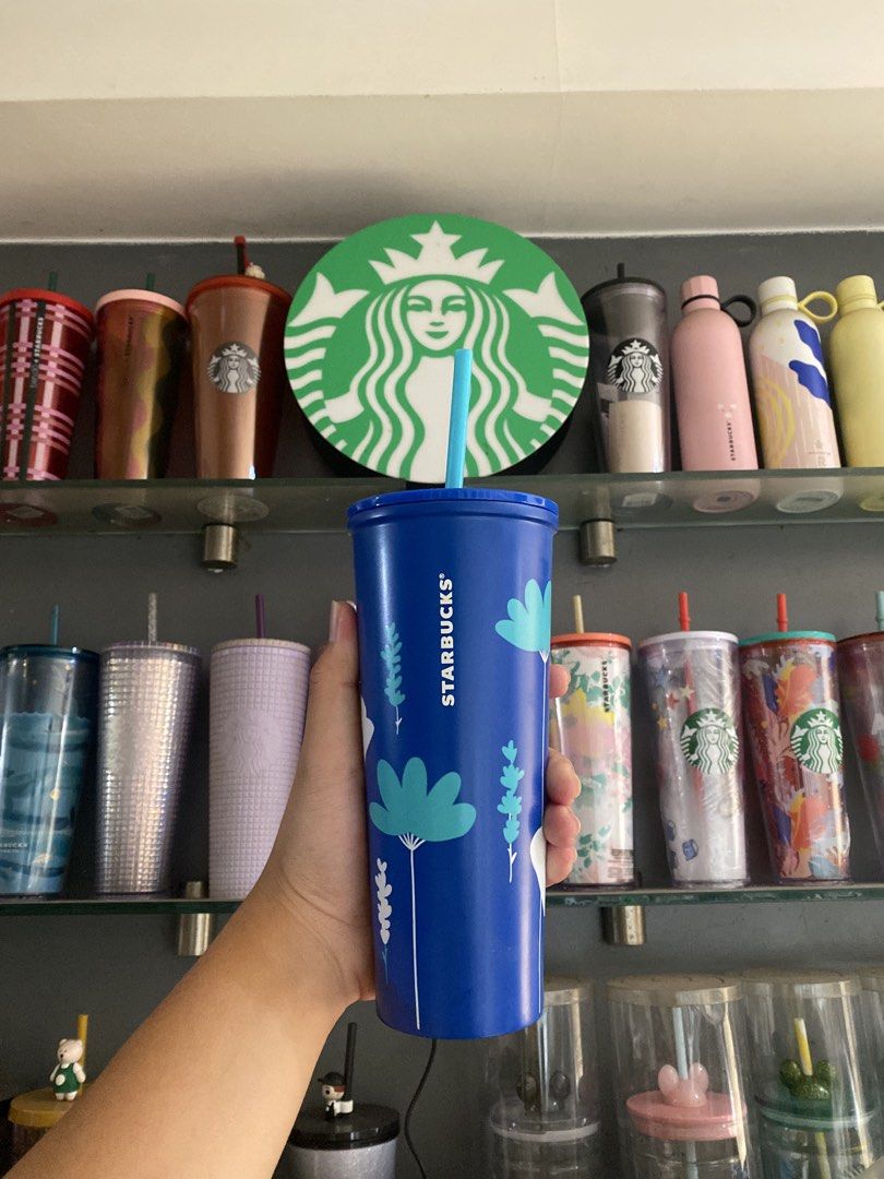 Starbucks blue Popoy flowers studded (Category: Water Bottles & Tumblers) located in Paranaque City. Submitted by jayisnottyfound (Listing ID 1466847182, Image 1). Description: • Primary Subject: A hand holds a tall, vibrant blue Starbucks seasonal cold cup tumbler in the foreground. • Background Setting: An multi-tiered glass shelf packed with a collection of colorful Starbucks merchandise (tumblers, water bottles, and mugs). • Focal Point: A la