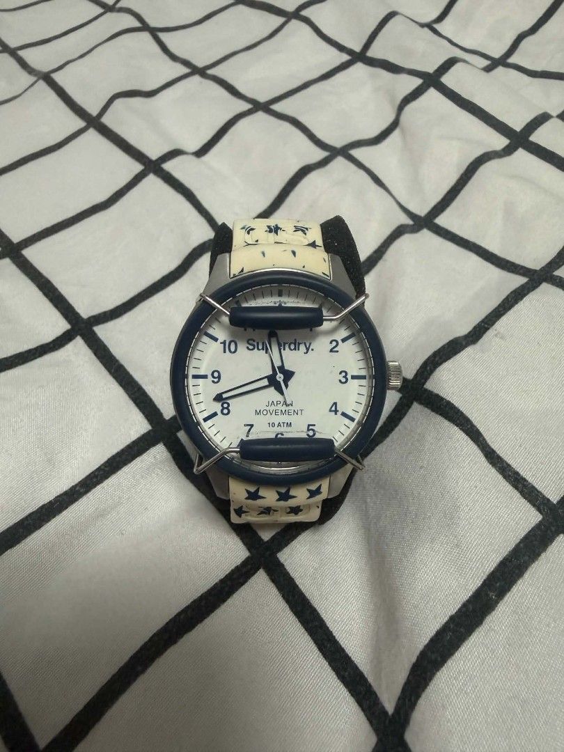 Superdry Japan Movement (Category: Women's Watches) located in Hermosa. Submitted by marywilma (Listing ID 1466864417, Image 1). Description: Superdry watch with white and blue star pattern strap. Blue bezel framing a white dial featuring bold numbers. Japan movement with 10 ATM water resistance. White deployment clasp stamped with Superdry logo. Stainless steel case back. Shows minor discoloration on the white strap links..