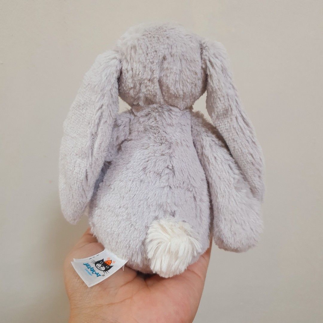 JELLYCAT Small Silver Bashful Bunny (Category: Plushies & Soft Toys) located in Quezon City. Submitted by mocha_mola (Listing ID 1421163417, Image 3). Description: Soft, gray Jellycat bunny stuffed toy. It has long floppy ears and a small white tail. The bunny has a small pink nose and a tag that reads "Jellycat London". The tag also indicates the item is made of polyester fibres and plastic pellets. The model number is Jelly7675..