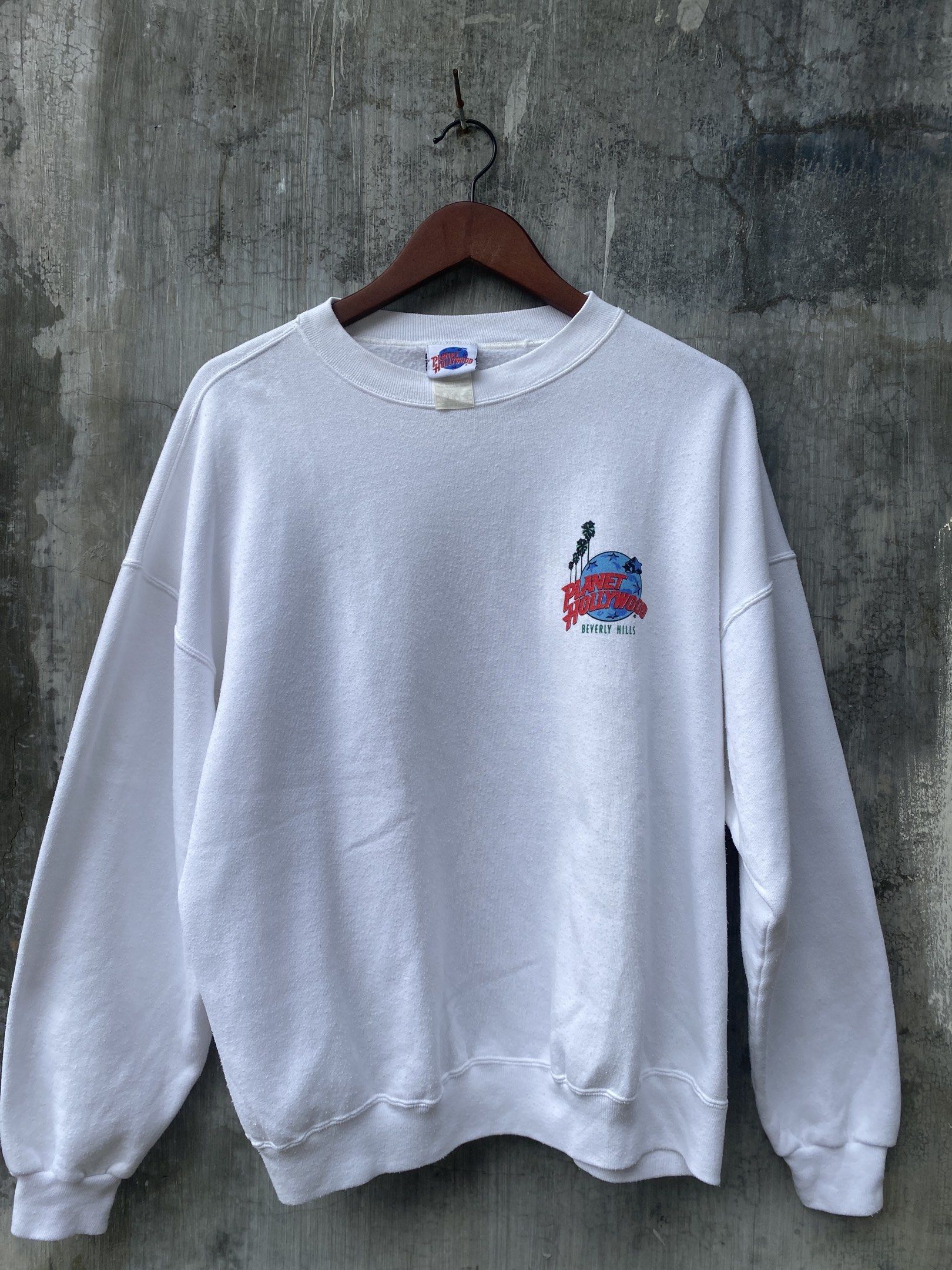 Planet Hollywood Beverly Hills Sweatshirt (Category: Coats, Jackets and Outerwear) located in San Pablo. Submitted by johnalmend72681 (Listing ID 1421342585, Image 1). Description: - White Planet Hollywood Beverly Hills sweatshirt - Features a small logo on the front - Large graphic on the back with a car and palm trees - Long sleeves - Crew neck - Made of soft material - Tag is visible.