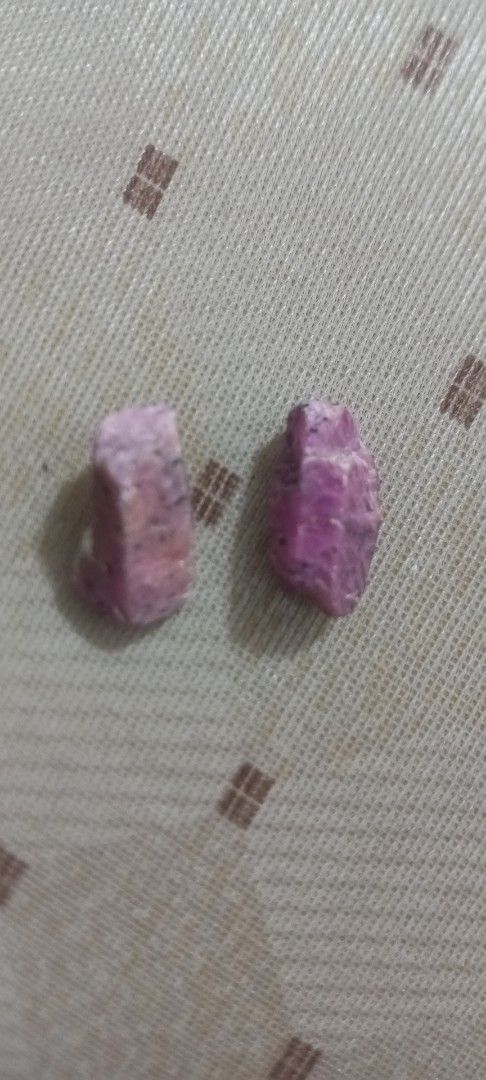 Raw Ruby Gemstones (Category: Precious Stones) located in Kota Kinabalu. Submitted by anas7389 (Listing ID 1421750485, Image 1). Description: - Two raw ruby gemstones - Pinkish-purple color - Natural, uncut form - Rough, textured surface - Shown on patterned fabric.
