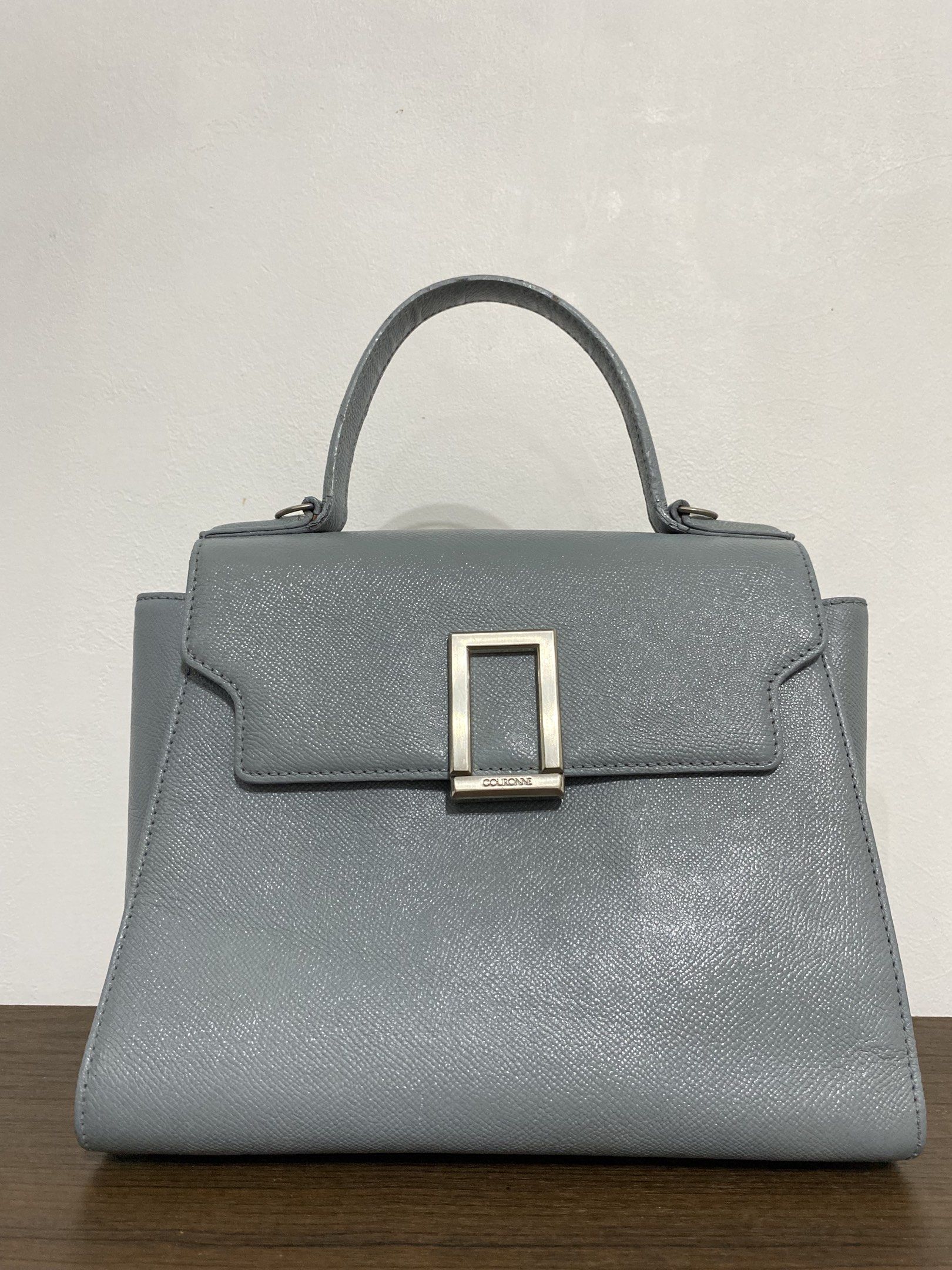 Couronne Handbag (Category: Luxury Bags & Wallets) located in Bulacan. Submitted by knklittlecloset (Listing ID 1421803343, Image 1). Description: Couronne handbag features a structured design with a top handle and a flap closure. It has a silver-tone metal detail on the front and a detachable shoulder strap. The interior includes a zippered pocket. The bag is in a light blue color. The bottom of the bag has four metal feet. Detachable strap and 