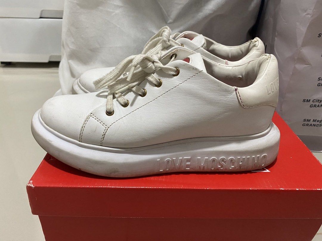 Love Moschino White Sneakers Size 38 (Category: Sneakers) located in Mandaluyong City. Submitted by getbounced (Listing ID 1421896849, Image 2). Description: White Love Moschino sneakers with heart details. Features a thick sole with "LOVE MOSCHINO" printed on the side. The size is 38. Includes the original box and a dust bag. The soles have heart-shaped patterns. The insole has "LOVE MOSCHINO" printed all over it..
