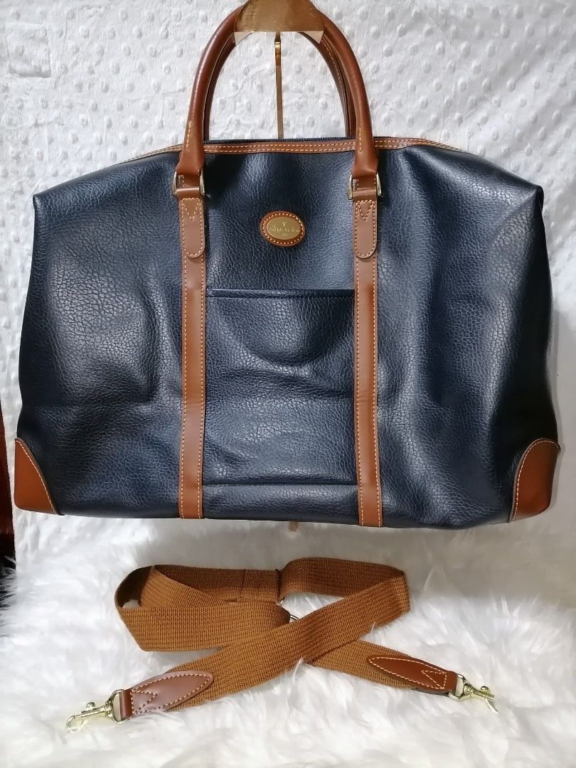 Rudolph Valentino Duffle Bag with Shoulder Strap (Category: Luxury Bags & Wallets) located in Butuan. Submitted by az-branded.items (Listing ID 1422024556, Image 2). Description: - Brand: Rudolph Valentino 1921 -By ACE Co. LTD. - Color: Navy blue with brown leather accents - Features: Detachable brown shoulder strap, front pocket, zippered closure - Interior: Black - Details: Brand name visible on bag and zipper -Size: W22" x L13"x Bottom 5.5".