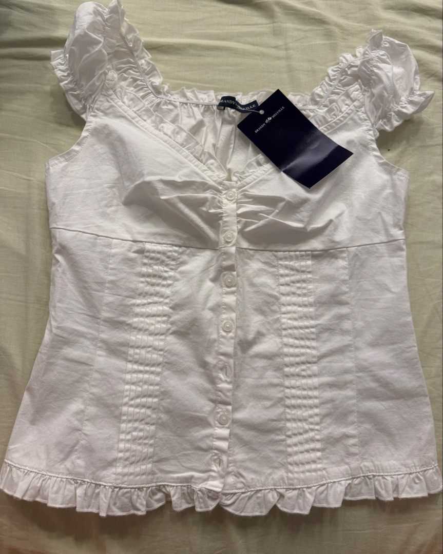 Brandy Melville White Elena Top (Category: Sleeveless) located in Bacacay. Submitted by hellogamer629 (Listing ID 1422351185, Image 1). Description: - Brand: Brandy Melville - Style: White top - Features: Button-down front, ruffled sleeves and hem, square neckline - Fit: Fitted silhouette - Condition: New with tag - Back: Plain with a seam.