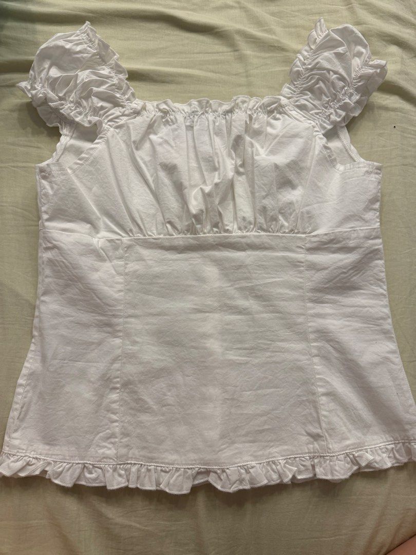 Brandy Melville White Elena Top (Category: Sleeveless) located in Bacacay. Submitted by hellogamer629 (Listing ID 1422351185, Image 2). Description: - Brand: Brandy Melville - Style: White top - Features: Button-down front, ruffled sleeves and hem, square neckline - Fit: Fitted silhouette - Condition: New with tag - Back: Plain with a seam.