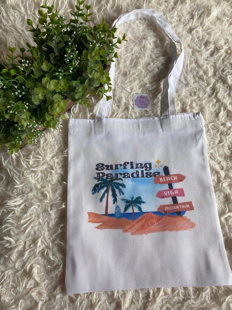 Surfing Paradise White Tote Bag (Category: Tote Bags) located in Cavite. Submitted by the-purple-cart (Listing ID 1422266813, Image 1). Description: - White tote bag - Dimension 12"x14" - 'Surfing Paradise' design - Features palm trees, surfboard, and signs - Two white handles - Durable canvas like material - Stylish and practical.