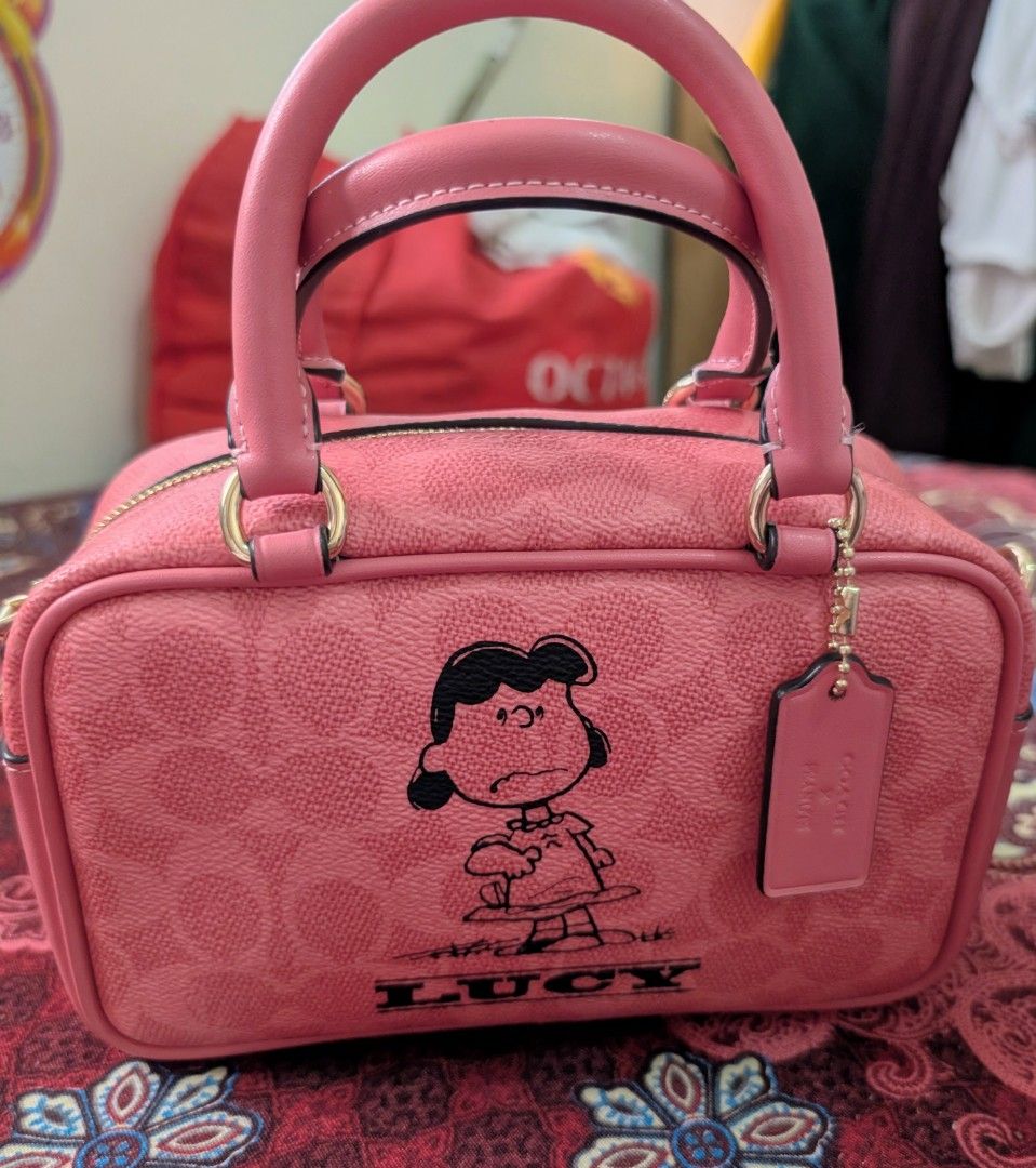 Coach x Peanuts Lucy Satchel (Category: Luxury Bags & Wallets) located in Rodriguez. Submitted by dessytrese13 (Listing ID 1419272199, Image 2). Description: - like new Coach satchel - Features Lucy from Peanuts - Stylish pink exterior - Includes a top handle and a detachable strap - Comes with a Coach hangtag - Showcases the iconic Coach logo pattern - Unused condition.