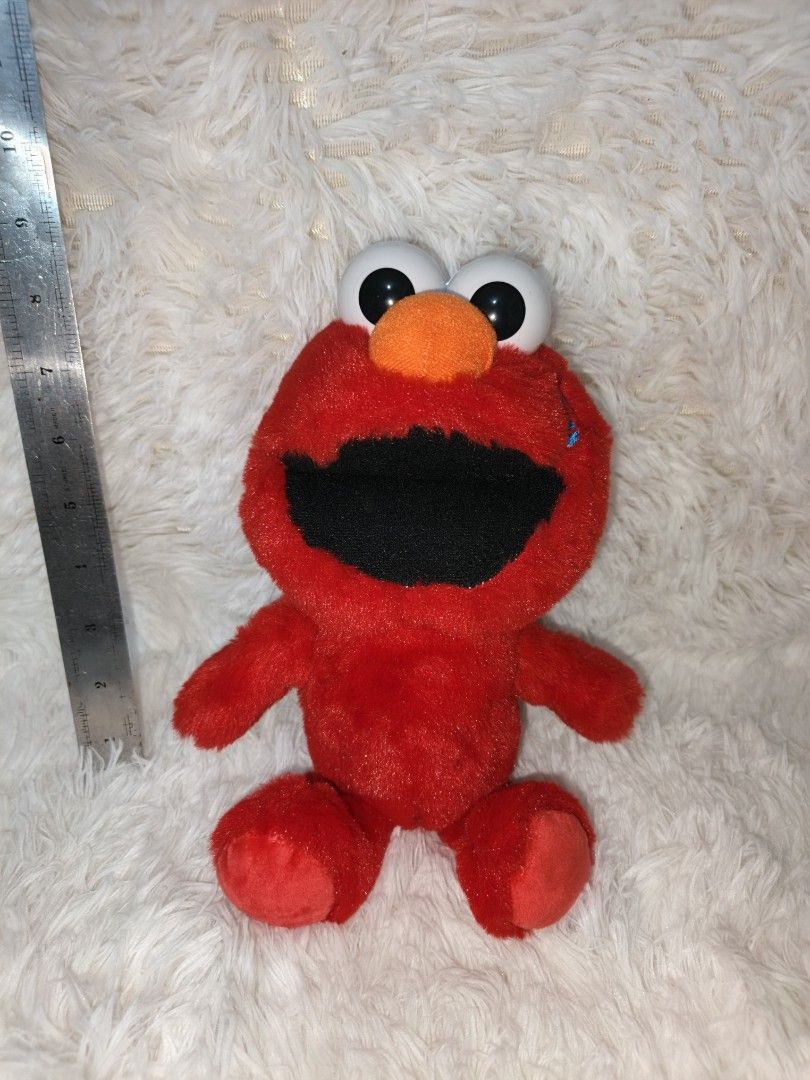Elmo Plush Toy (Category: Plushies & Soft Toys) located in Marikina City. Submitted by andreah_16 (Listing ID 1423410217, Image 3). Description: Red Elmo plush toy. It has big white eyes, an orange nose, and a black mouth. The toy is made of soft, red plush material. It has arms and legs. The toy is sitting on a white, fluffy surface. A ruler is placed next to the toy for size reference. paki check nalang po sa ruler yung size 📍loc : sto. niño m