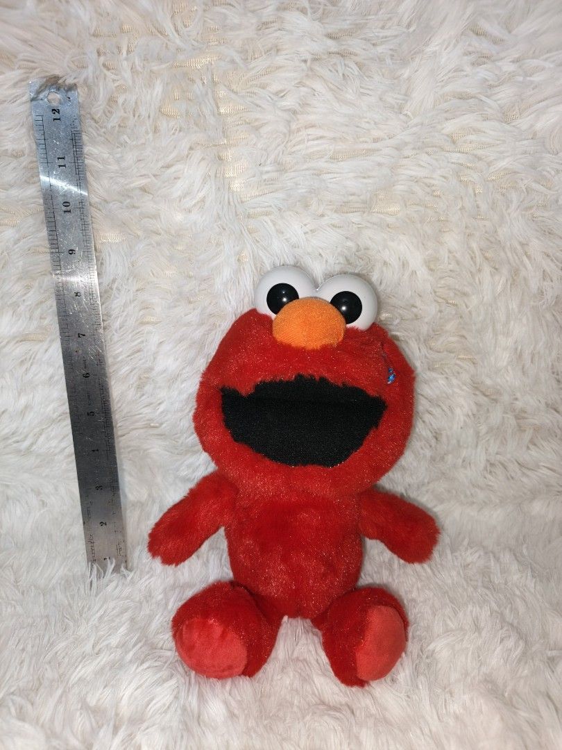 Elmo Plush Toy (Category: Plushies & Soft Toys) located in Marikina City. Submitted by andreah_16 (Listing ID 1423410217, Image 2). Description: Red Elmo plush toy. It has big white eyes, an orange nose, and a black mouth. The toy is made of soft, red plush material. It has arms and legs. The toy is sitting on a white, fluffy surface. A ruler is placed next to the toy for size reference. paki check nalang po sa ruler yung size 📍loc : sto. niño m