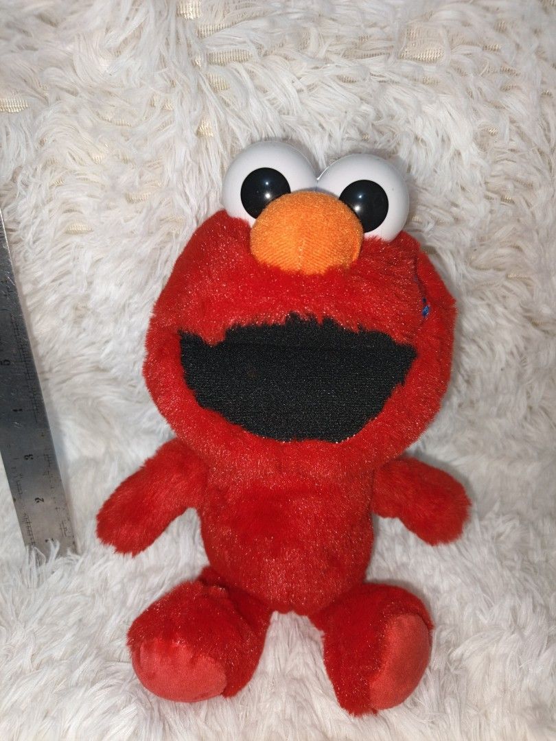 Elmo Plush Toy (Category: Plushies & Soft Toys) located in Marikina City. Submitted by andreah_16 (Listing ID 1423410217, Image 1). Description: Red Elmo plush toy. It has big white eyes, an orange nose, and a black mouth. The toy is made of soft, red plush material. It has arms and legs. The toy is sitting on a white, fluffy surface. A ruler is placed next to the toy for size reference. paki check nalang po sa ruler yung size 📍loc : sto. niño m