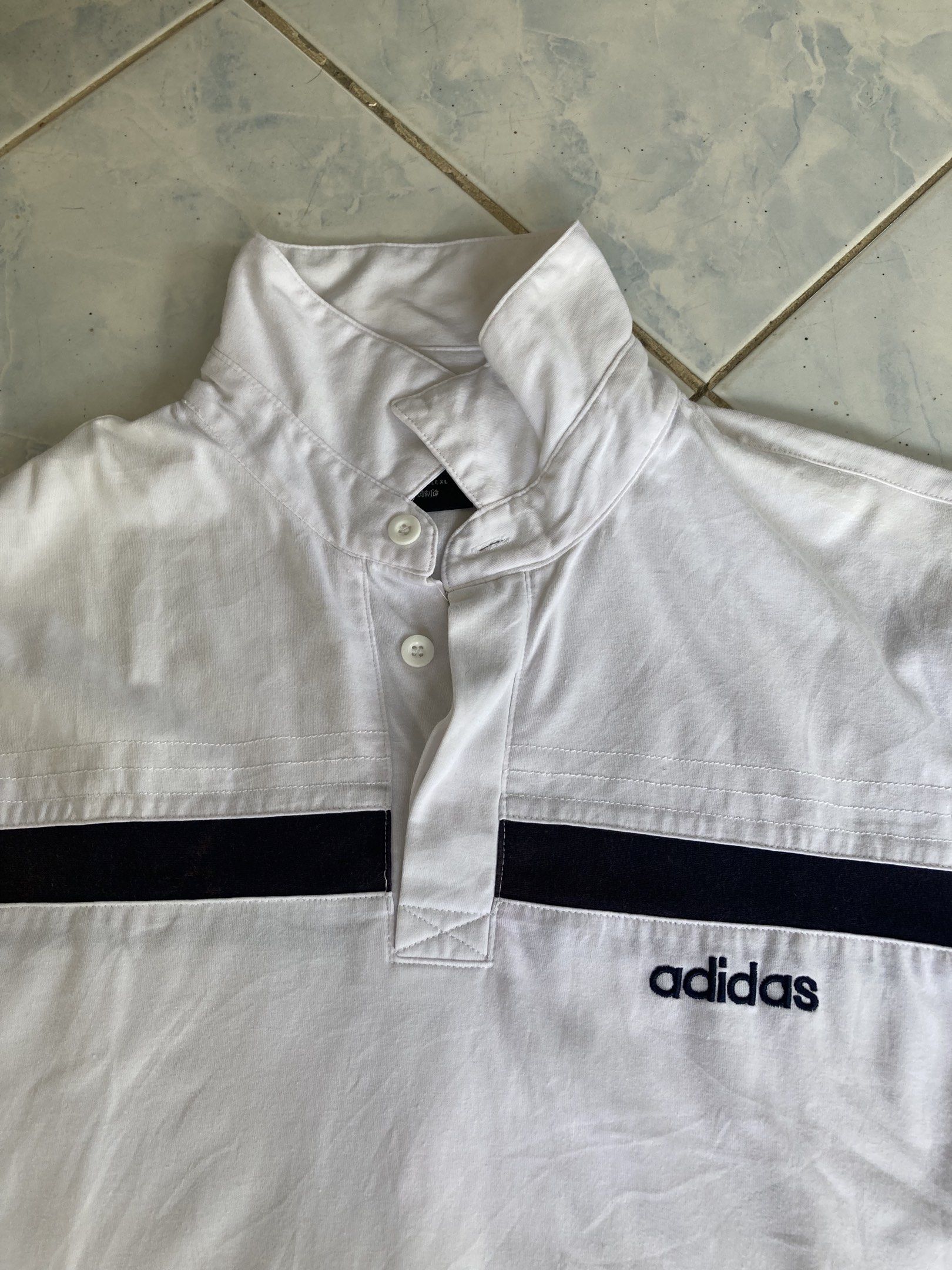 Adidas White Polo Shirt (Category: Tshirts & Polo Shirts ) located in Tarlac. Submitted by everthrift (Listing ID 1419776860, Image 2). Description: - White Adidas polo shirt - Black stripes across chest and back - High collar - Adidas logo on chest - Short sleeves - Button-up neckline - Minor discoloration on back stripe.
