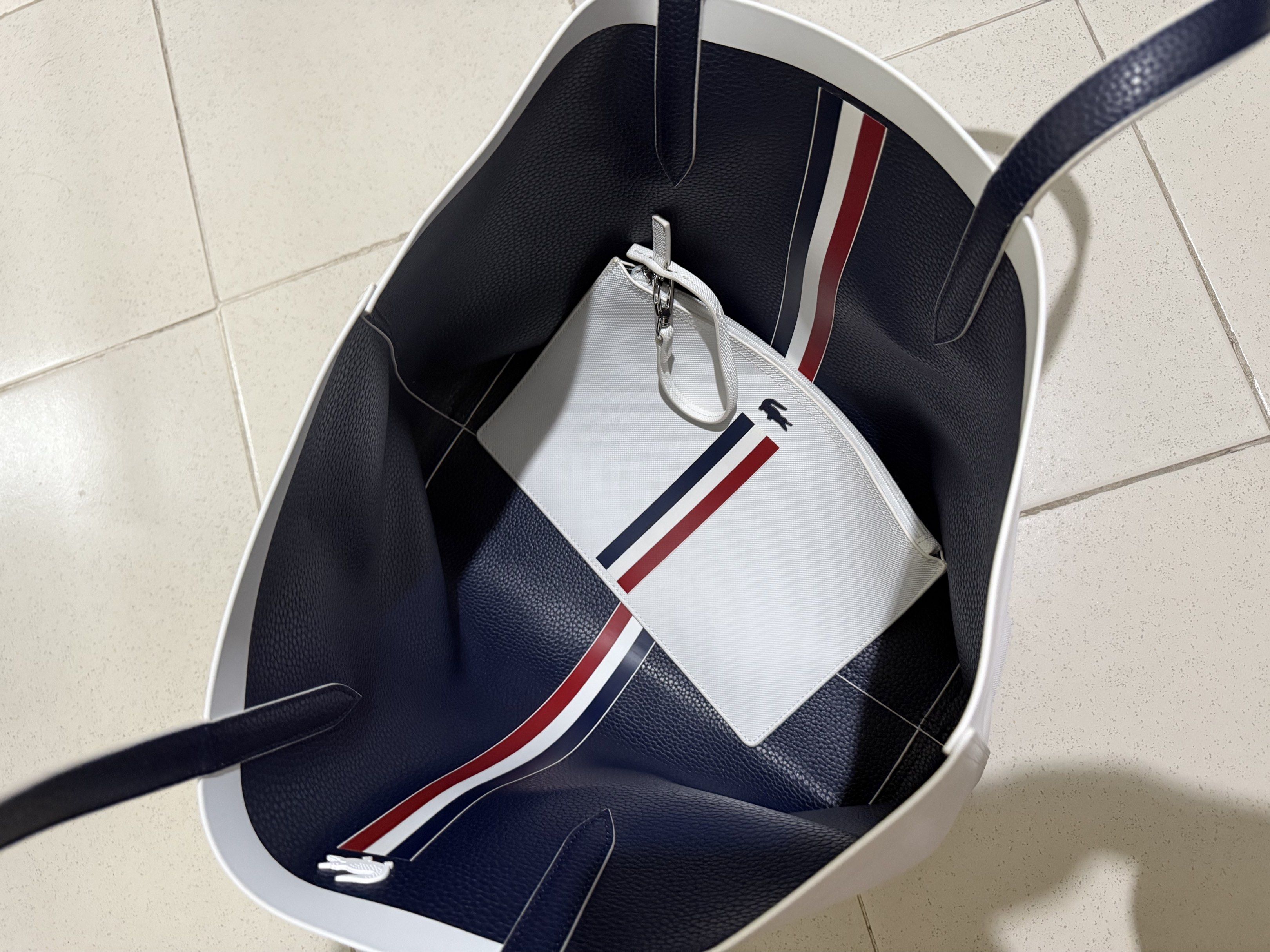 Lacoste Reversible womens large tote bag (Category: Luxury Bags & Wallets) located in Apalit. Submitted by xoxo.serena (Listing ID 1420009421, Image 2). Description: Brand: Lacoste • Color: White with classic tricolor stripe (navy/red) • Includes: Tote bag + original matching pouch • Material: Textured synthetic/leather-look material • Condition: Very good condition (no visible stains or damage) • Spacious interior, perfect for daily use, school,