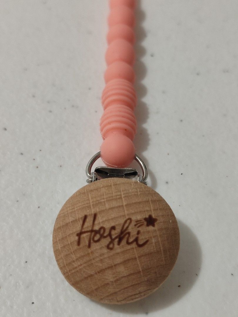 Hoshi Silicon String Pacifier Clip (Category: Soothers & Pacifiers) located in Marikina City. Submitted by surplusbuffet (Listing ID 1420295402, Image 2). Description: Hoshi pacifier clip features a wooden disc with the brand name and a star. It has a pink silicone beaded chain and a clip to attach to clothing. The clip is designed to securely hold a pacifier, preventing it from getting lost or dropped. The clip is made of wood and metal..