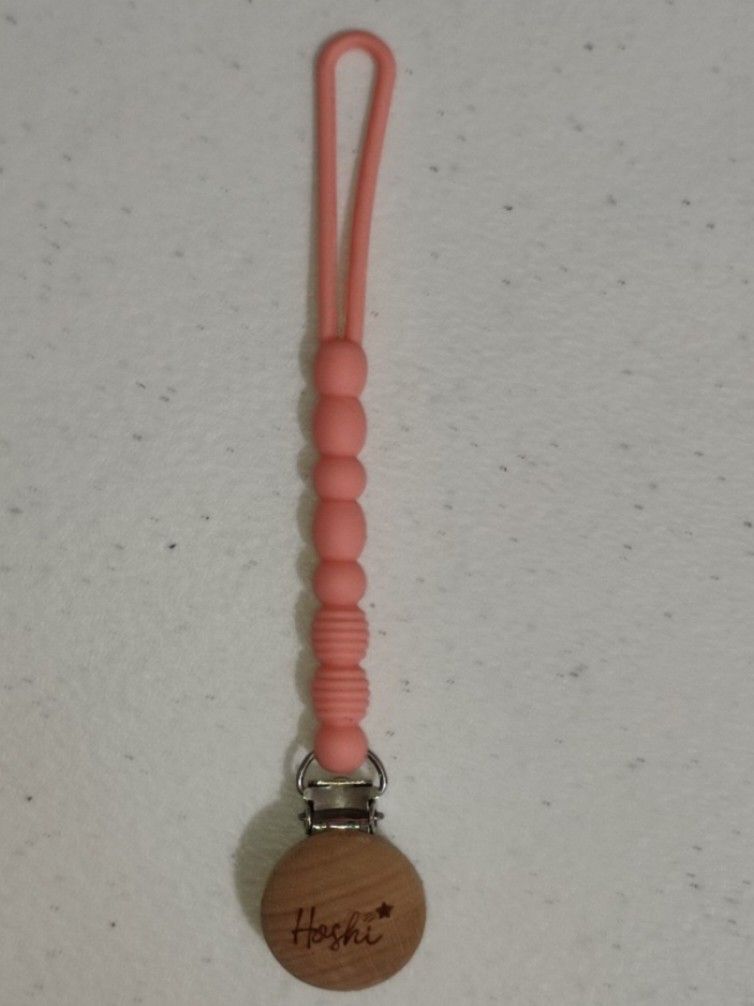 Hoshi Silicon String Pacifier Clip (Category: Soothers & Pacifiers) located in Marikina City. Submitted by surplusbuffet (Listing ID 1420295402, Image 3). Description: Hoshi pacifier clip features a wooden disc with the brand name and a star. It has a pink silicone beaded chain and a clip to attach to clothing. The clip is designed to securely hold a pacifier, preventing it from getting lost or dropped. The clip is made of wood and metal..