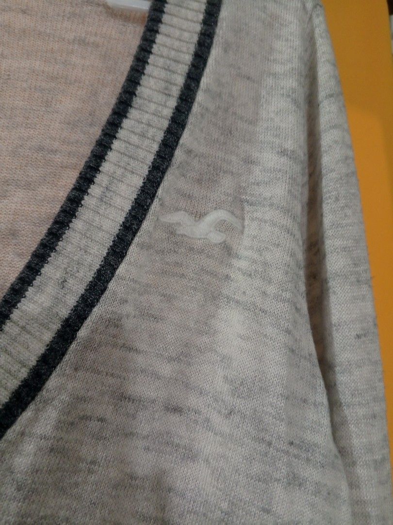 y2k Hollister V-Neck Sweater (Category: Coats, Jackets and Outerwear) located in Lucena. Submitted by minos_world (Listing ID 1425980723, Image 2). Description: Hollister V-neck sweater in light gray with navy blue trim. Features the Hollister logo on the chest. The size is Small. The sweater has long sleeves with navy blue trim at the cuffs. The tag is visible, indicating the brand and size..