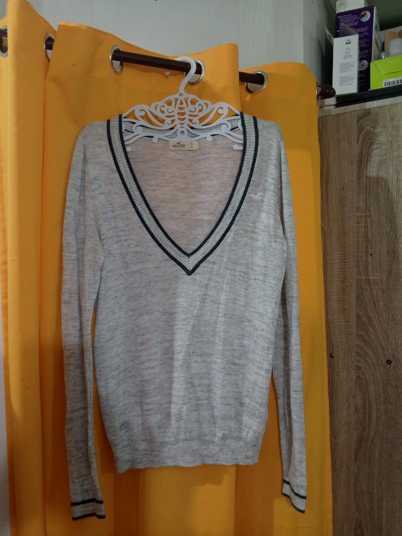 y2k Hollister V-Neck Sweater (Category: Coats, Jackets and Outerwear) located in Lucena. Submitted by minos_world (Listing ID 1425980723, Image 1). Description: Hollister V-neck sweater in light gray with navy blue trim. Features the Hollister logo on the chest. The size is Small. The sweater has long sleeves with navy blue trim at the cuffs. The tag is visible, indicating the brand and size..