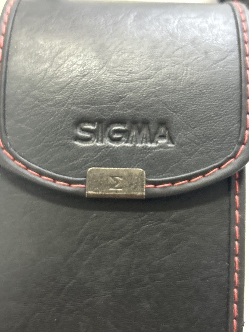 Sigma Camera Case with Strap (Category: Camera Bags & Carriers) located in Bulacan. Submitted by ourdailybreads (Listing ID 1425841402, Image 2). Description: Black Sigma camera case with a red stitched trim. Includes a black strap for carrying. The case has a rectangular shape with a flap closure. The Sigma logo is visible on the front..