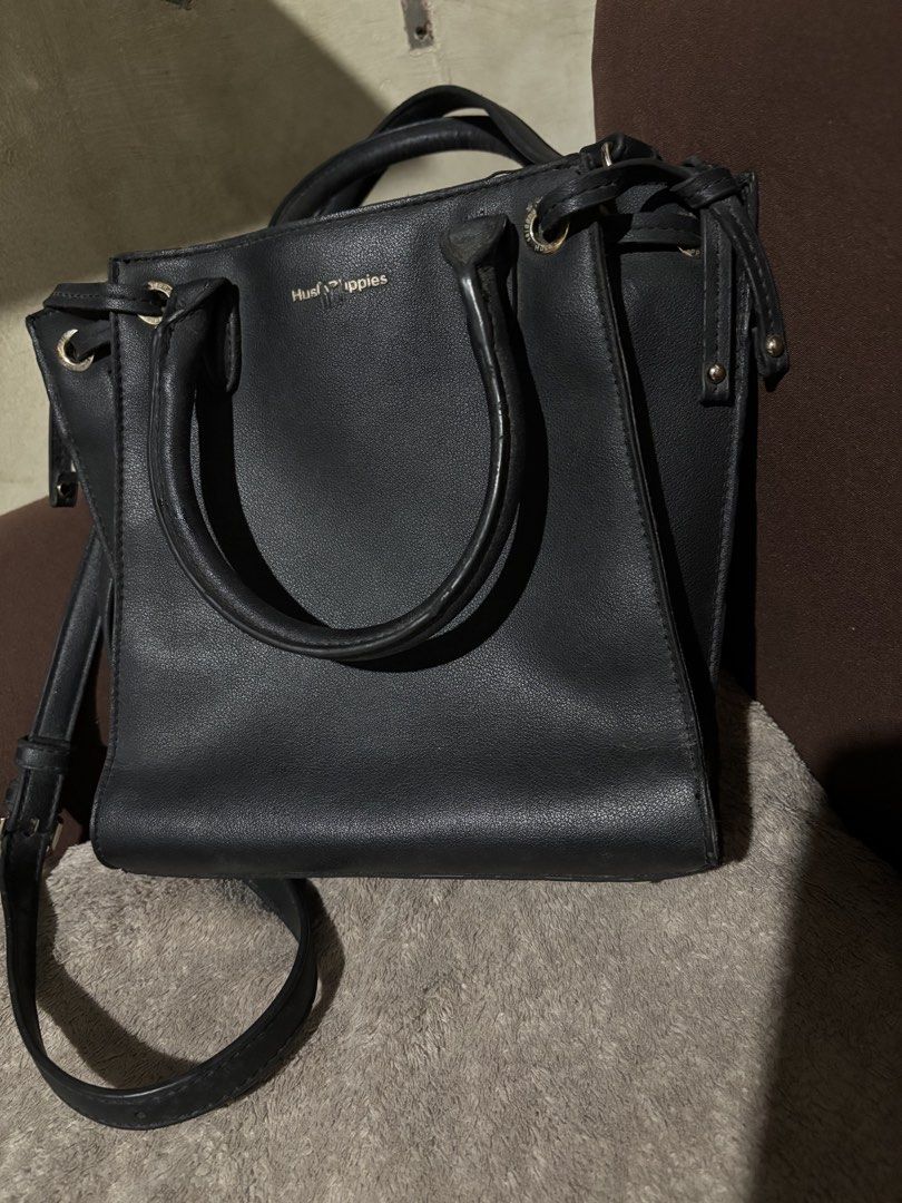Hush Puppies Black Handbag (Category: Luxury Bags & Wallets) located in Batangas. Submitted by mylenevillegas-21 (Listing ID 1426150202, Image 2). Description: Black Hush Puppies handbag with top handles and a detachable shoulder strap. Features a magnetic snap closure and a zippered interior pocket. The bag has a structured shape with metal feet on the bottom. The interior is lined with fabric and includes a brand label. The exterior has a subtl