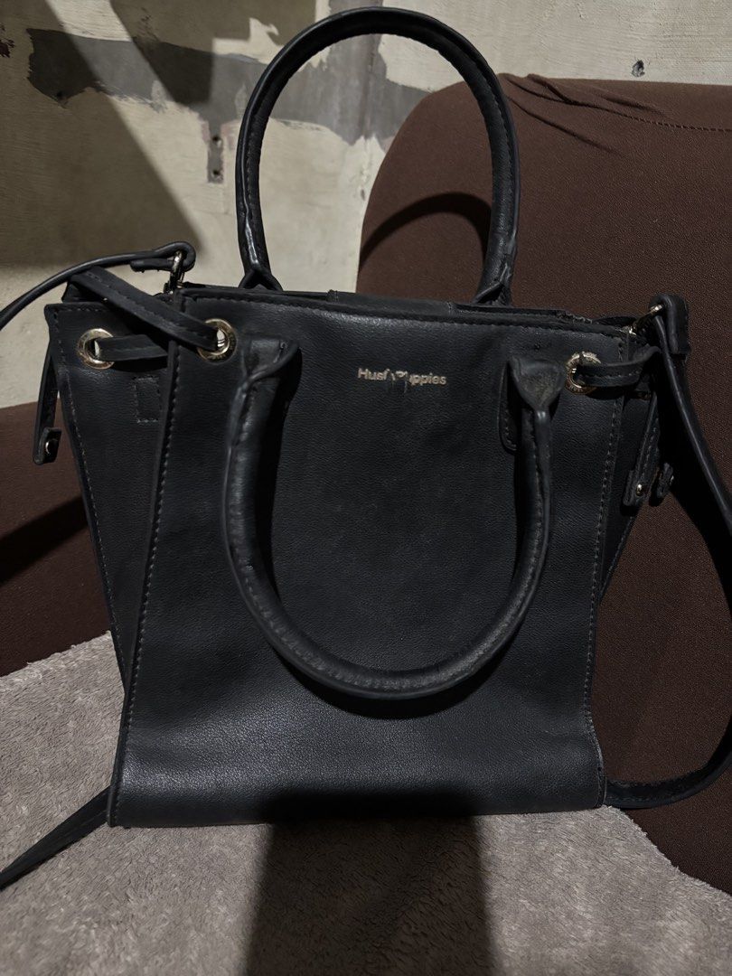 Hush Puppies Black Handbag (Category: Luxury Bags & Wallets) located in Batangas. Submitted by mylenevillegas-21 (Listing ID 1426150202, Image 1). Description: Black Hush Puppies handbag with top handles and a detachable shoulder strap. Features a magnetic snap closure and a zippered interior pocket. The bag has a structured shape with metal feet on the bottom. The interior is lined with fabric and includes a brand label. The exterior has a subtl
