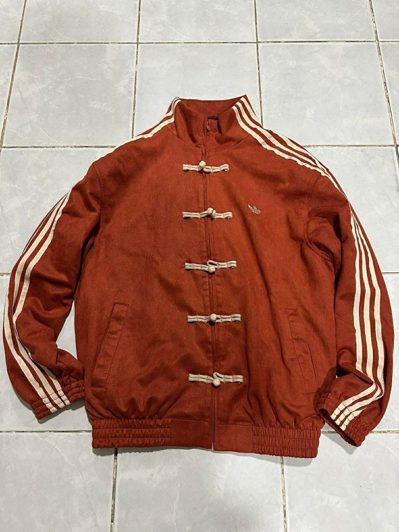 Adidas Track Jacket with Chinese Knot Buttons (Category: Coats, Jackets and Outerwear) located in Manila. Submitted by ukaygandamo (Listing ID 1426199350, Image 1). Description: Adidas track jacket features a unique design with Chinese knot buttons. It has the classic Adidas three stripes on the sleeves and the Adidas logo on the chest. The jacket has a mandarin collar and a zipper closure. The color is a reddish-orange. The jacket has a ribbed h