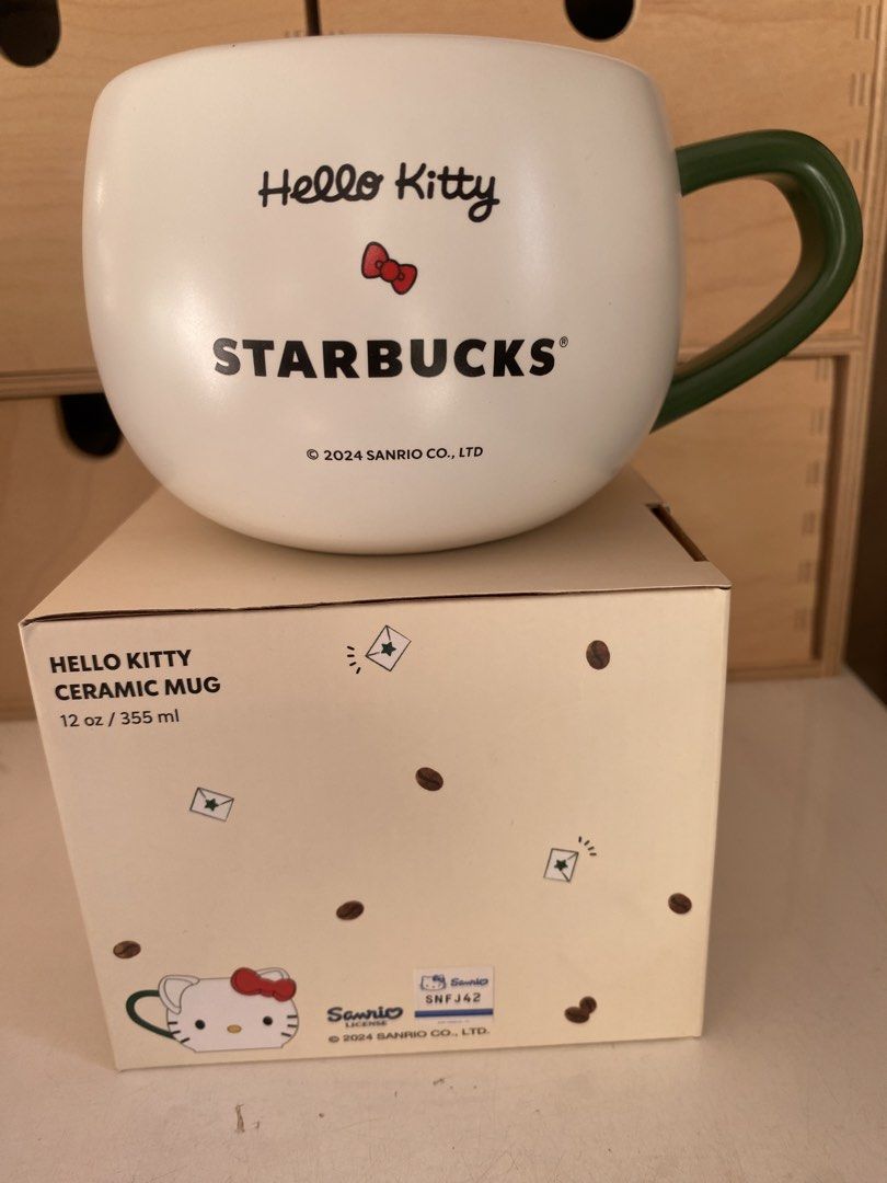 Hello Kitty Starbucks Ceramic Mug (Category: Coffee & Tea Tableware) located in Pasay City. Submitted by trendystash (Listing ID 1426485084, Image 2). Description: Brand new with box Kitty ceramic mug is a collaboration with Starbucks. It features a Kitty design with a red bow and a green handle. The mug has the Starbucks logo and is marked as 12 oz / 355 mL. It is microwave and dishwasher safe..