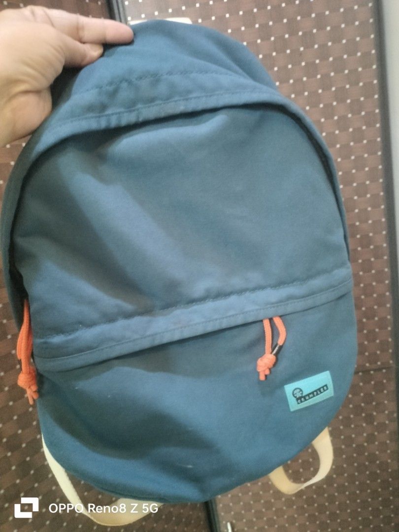 Crumpler Backpack (Category: Backpacks) located in Tanza. Submitted by ireennee (Listing ID 1427516253, Image 1). Description: Crumpler backpack features a teal-colored exterior with orange drawstring details. It has a main compartment and a smaller front pocket. The Crumpler logo is visible on a light blue patch. The backpack has a simple, rounded design with a top handle and adjustable straps. The straps are a light beige color..