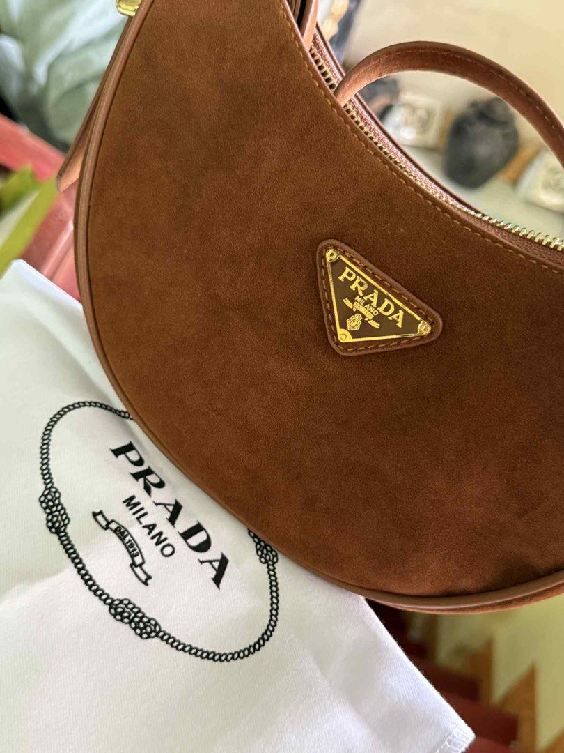 Prada Arque Suede Bag (Category: Luxury Bags & Wallets) located in Cabuyao City. Submitted by girliecloset202434 (Listing ID 1427527811, Image 2). Description: Brown suede Prada hobo bag. Features a gold-tone zipper and a triangular Prada logo on the front. The interior has a Prada label. Includes a Prada dust bag. The bag has a curved shape and a single shoulder strap..