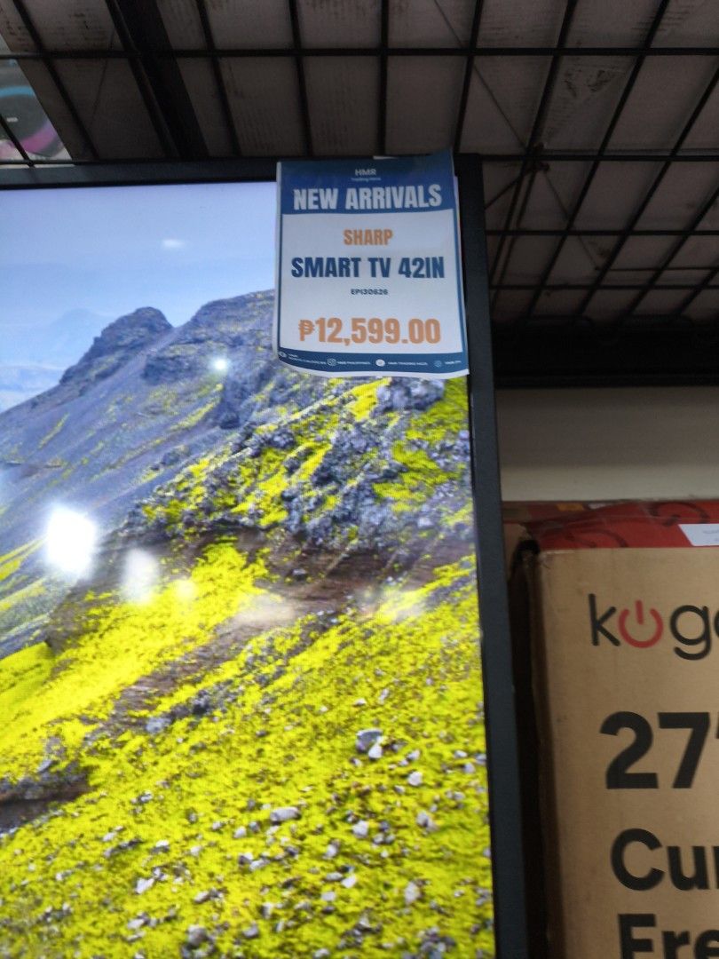 Sharp Smart TV 42in (Category: TV) located in Manila. Submitted by hmrgrace5. (Listing ID 1427493078, Image 2). Description: Sharp Smart TV with a 42-inch screen. The TV displays a vibrant landscape scene. It has a black frame and comes with a stand. The price tag indicates a price of ₱12,599.00. The TV is a new arrival..